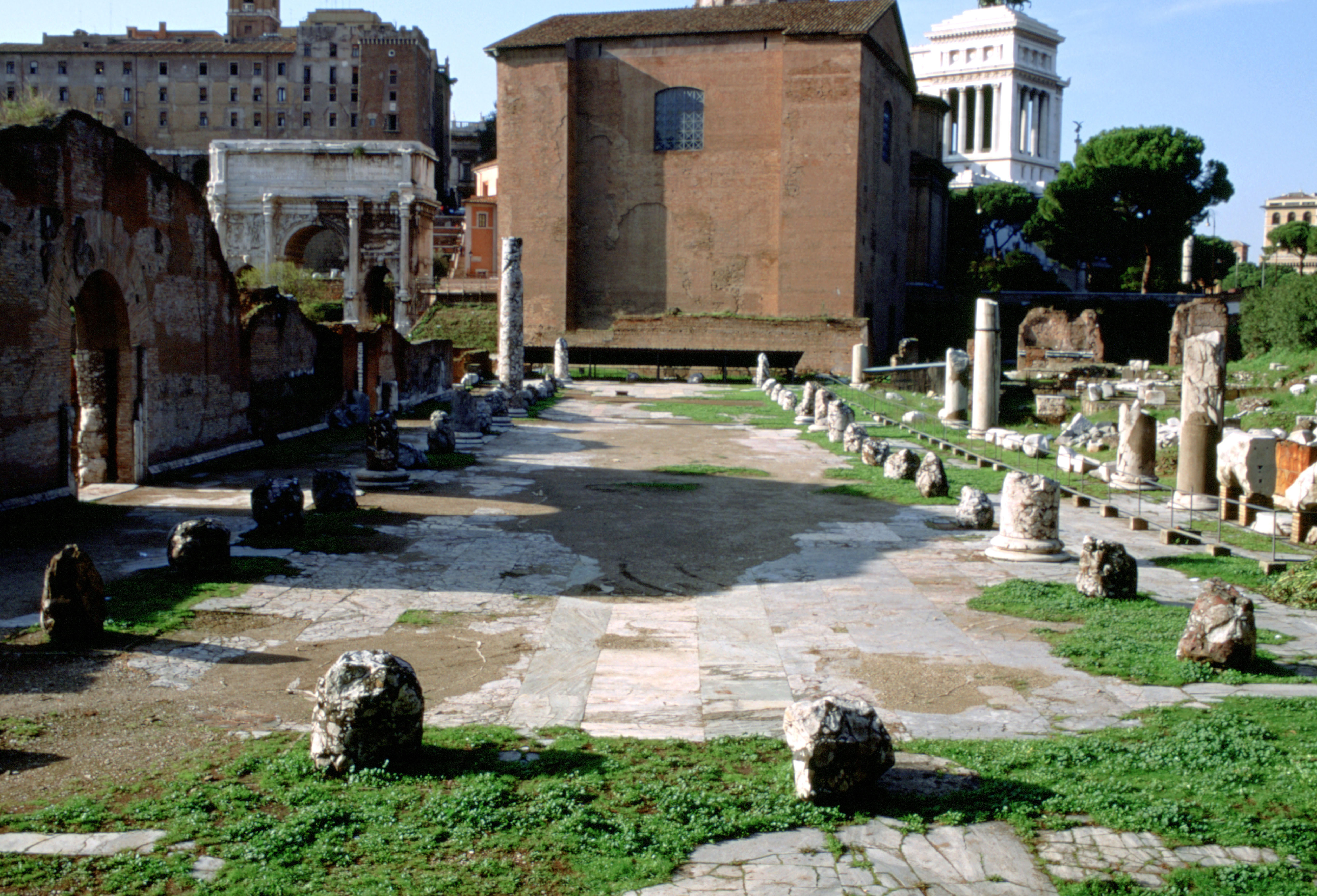 A long paved area with the remains of columns aroud the sides. At the end of the paved area is a building in red brick.
