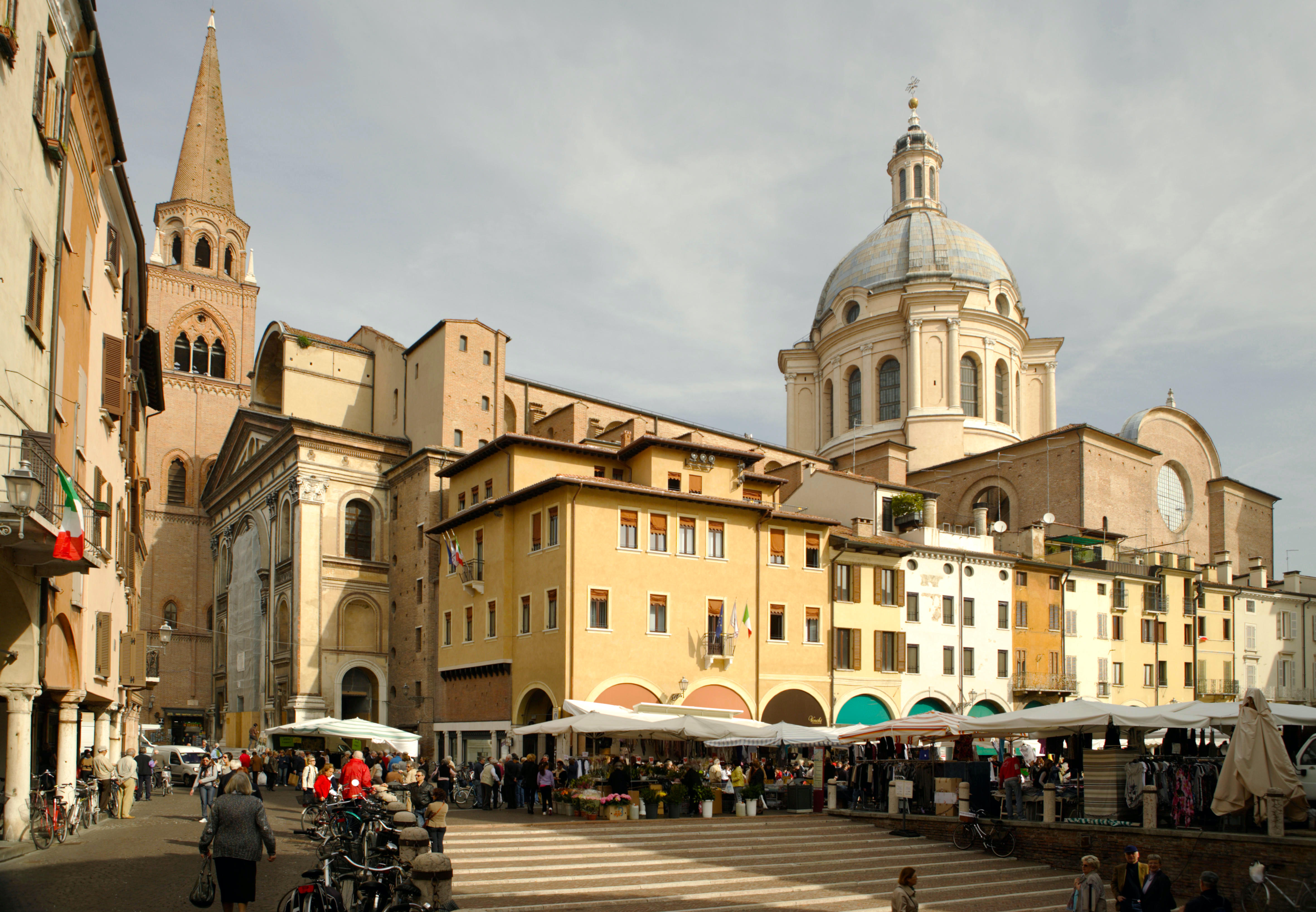 A photo of the town square in Mantua, Italy. There is a tall church dome and a spire. There are municipal buildings with an italian flags hanging from the window. There are market stalls with shoppers gathered round and groups of people chatting. On the left there is a covered walkway.